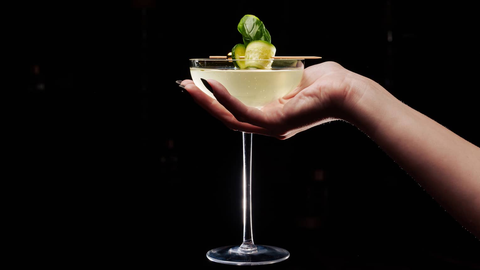 A hand holding a coupe cocktail glass filled with a yellow liquid and garnished with a curled piece of cucumber on a toothpick