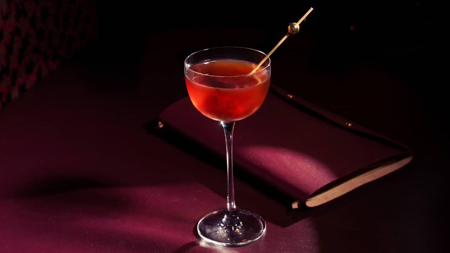 A bright red beverage in a tall stemmed glass garnished with a long gold stirrer