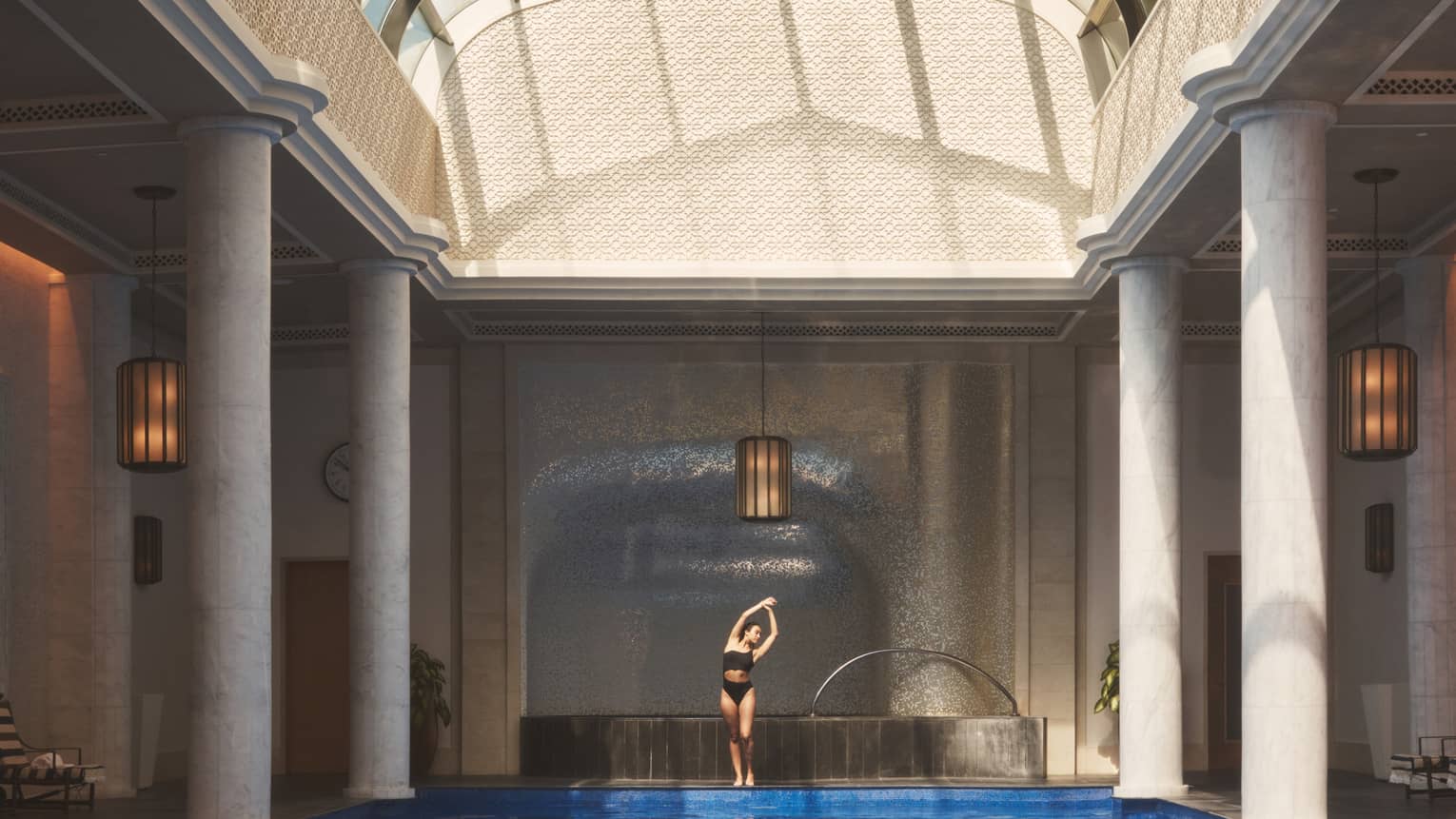 Person in swimsuit standing and stretching with arms overhead beside indoor pool flanked with white pillars under vaulted ceiling