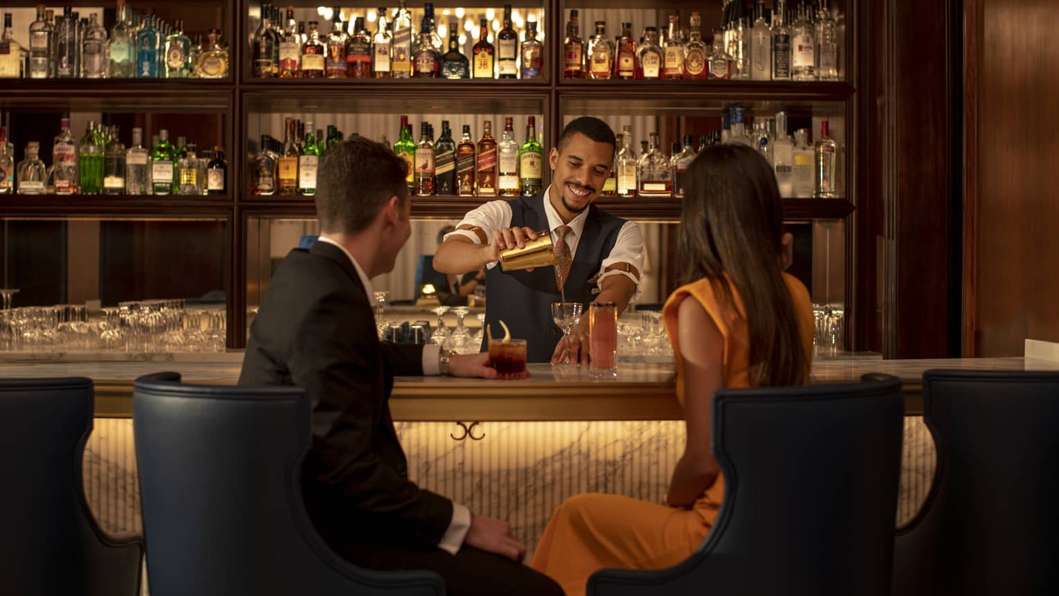 Man and woman sit at bar, bartender pours cocktail
