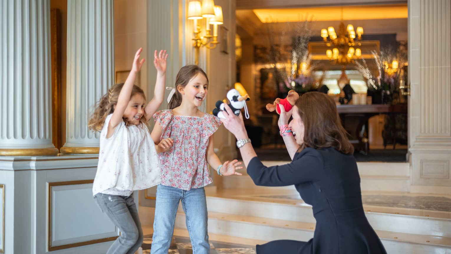 Concierge crouched, holding two stuffed animals toward excited, smiling girls