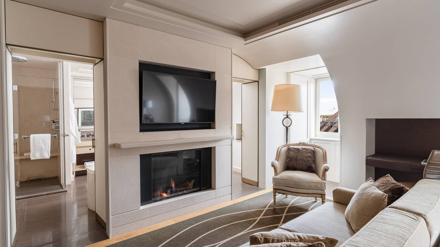 Luxury hotel room with a sofa, modern fireplace, wall-mounted TV, armchair near a window and view of an adjacent bathroom