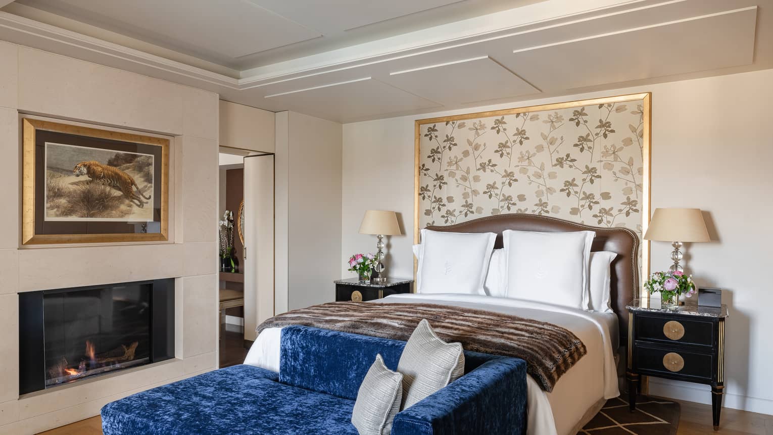 Luxury hotel bedroom with a king bed, floral headboard, elegant brown bedframe, plush blue bench, fireplace and bedside tables topped with lamps and flowers