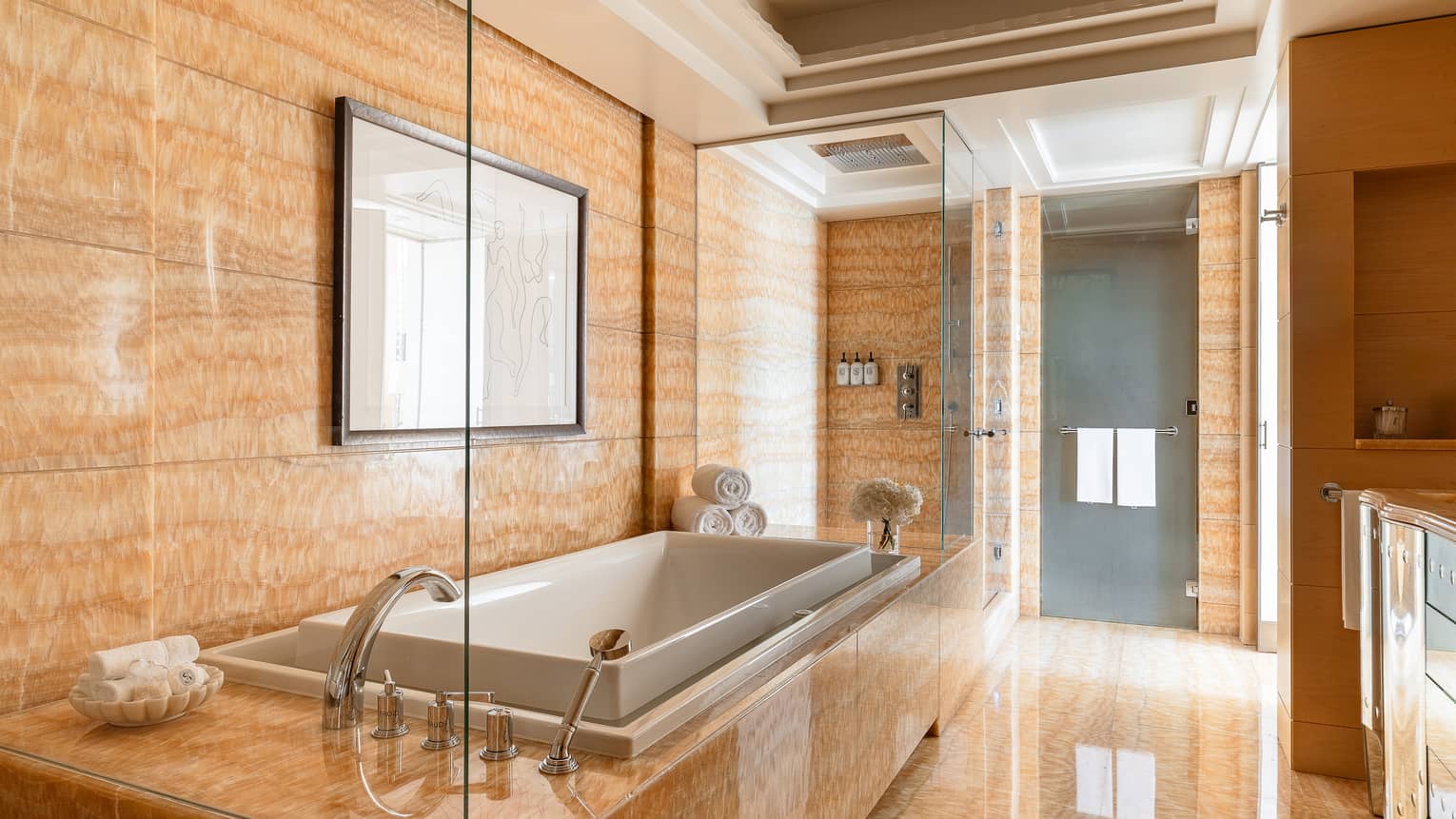 Presidential suite luxury bathroom with warm marble tiles, a large built-in bathtub, glass-enclosed rain shower and rolled towels placed near a vase of flowers