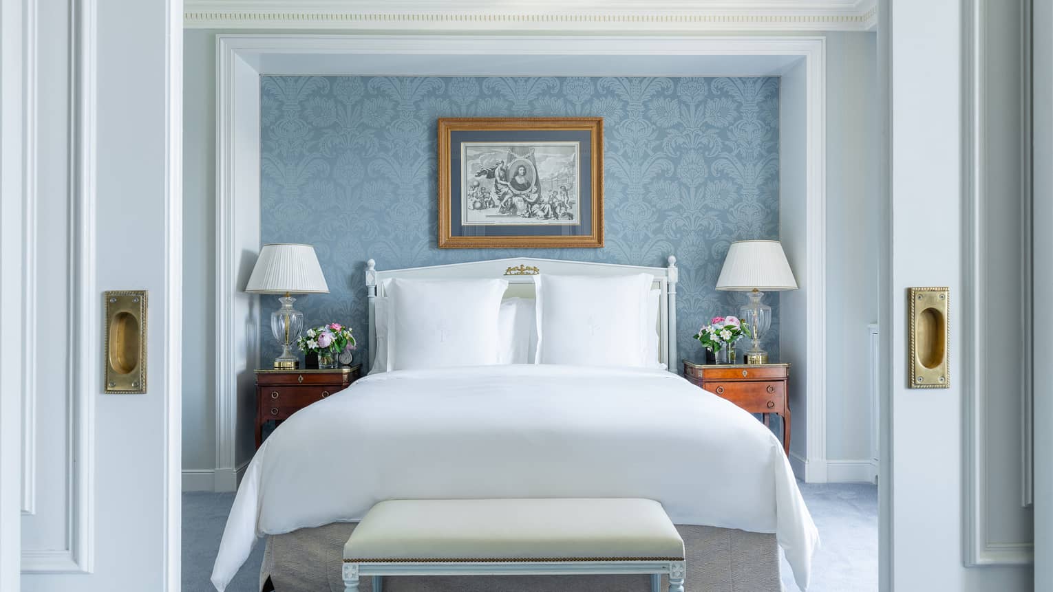 Luxury hotel room with soft blue undertones, featuring a king bed, classic wooden bedside tables with lamps and flowers, floral wallpaper and framed artwork