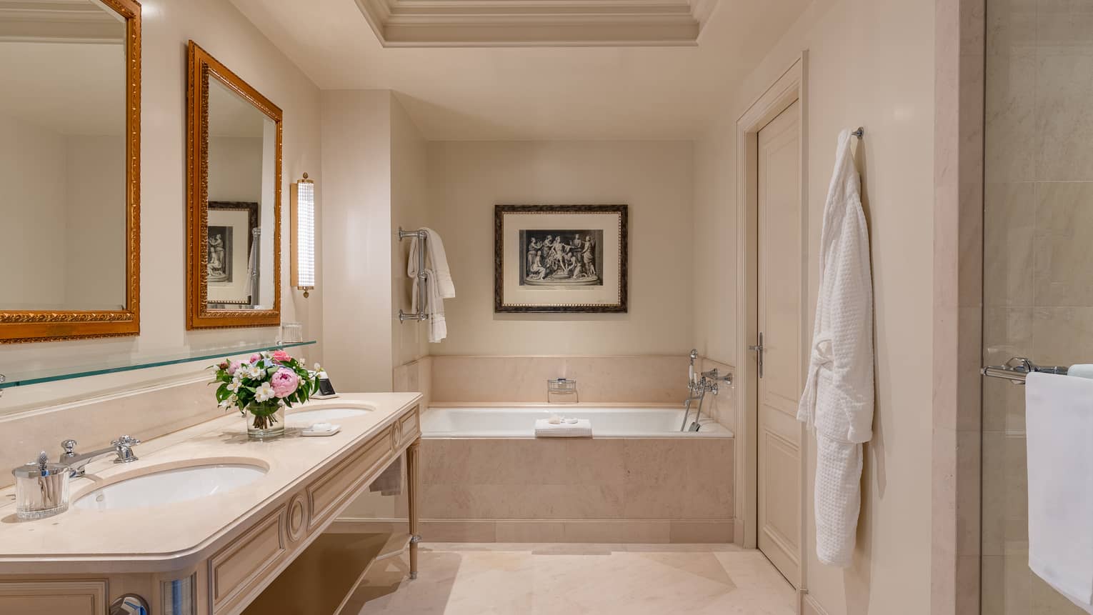 Luxury hotel bathroom with a double sink vanity, gold-framed mirrors, bathrobe hanging on the wall and a bathtub adorned by elegant artwork