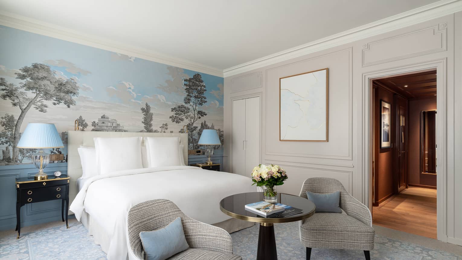 Hotel guest room with white king bed, blue mural accent wall, two grey arm chairs and round table at the foot of the bed, and small hallway