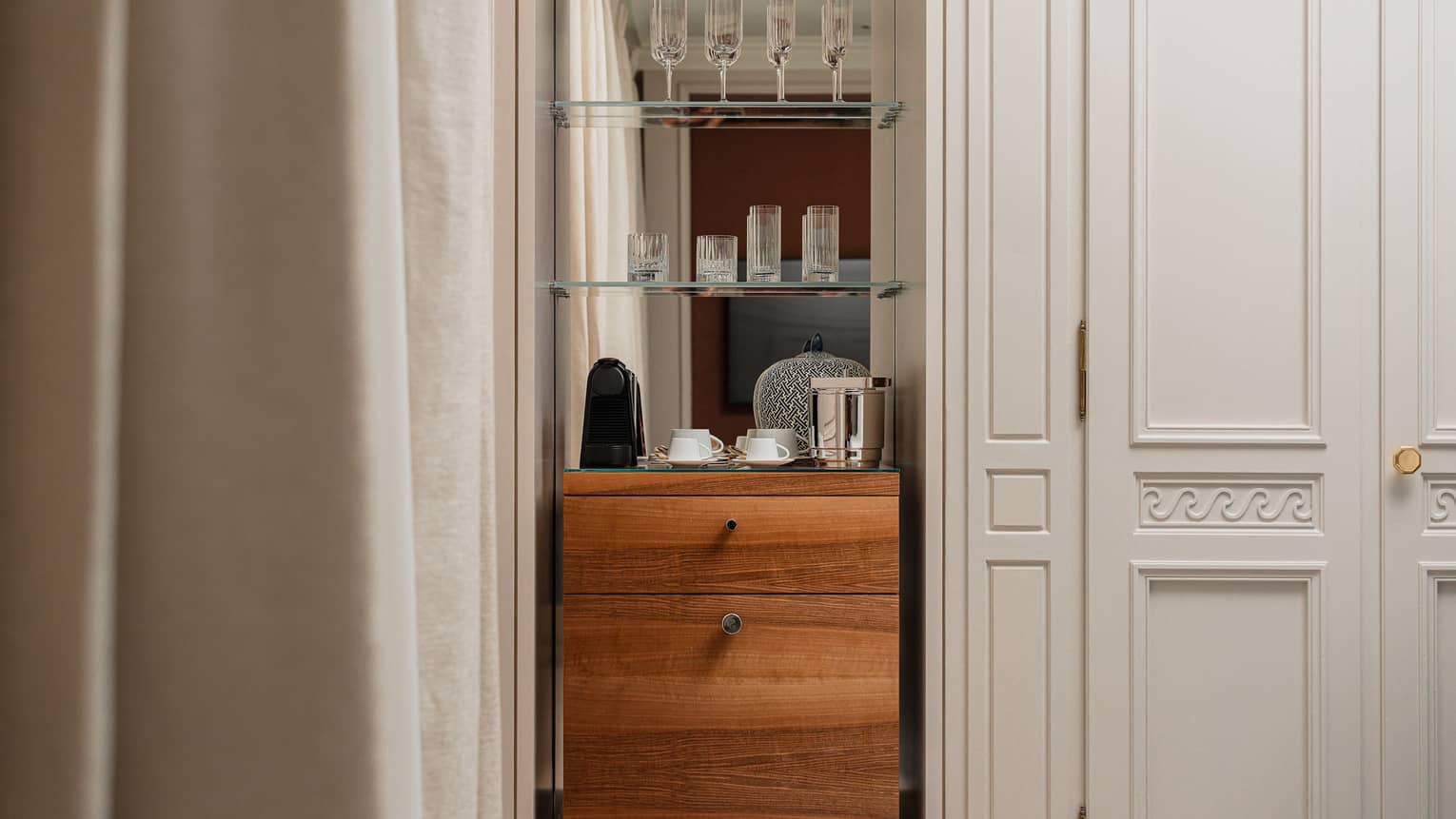 A small beverage counter and overhead glass shelves holding glasses in a hotel room
