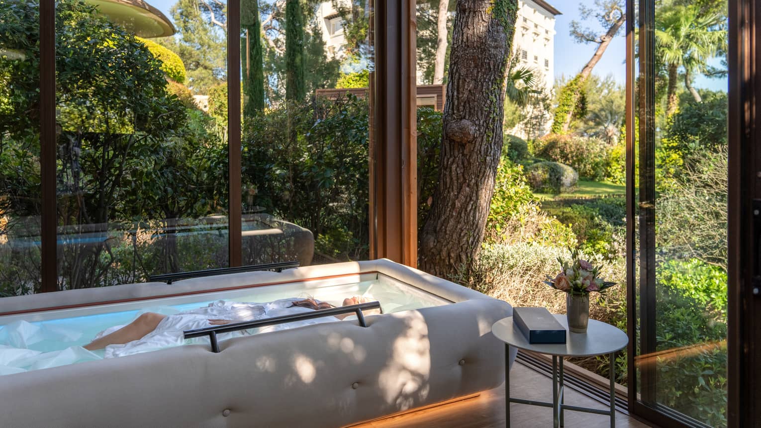 A serene glass-enclosed spa space with a small, raised, oblong pool. Trees and greenery are visible through the glass walls and surround the space.
