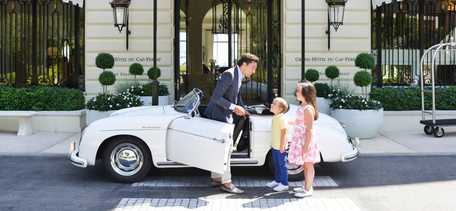 Concierge talking to boy in yellow shirt, girl in pink sundress beside white vintage convertible, in front of hotel