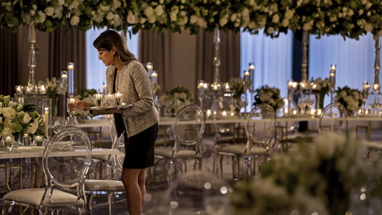A member of the hotel setting up for a wedding by placing candles on long elegant tables that have flowers as center pieces.