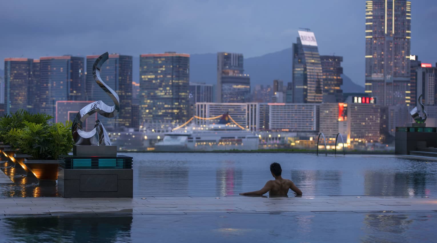 Silhouette of back of man wading in infinity swimming pool, looking out at Hong Kong harbour at dusk