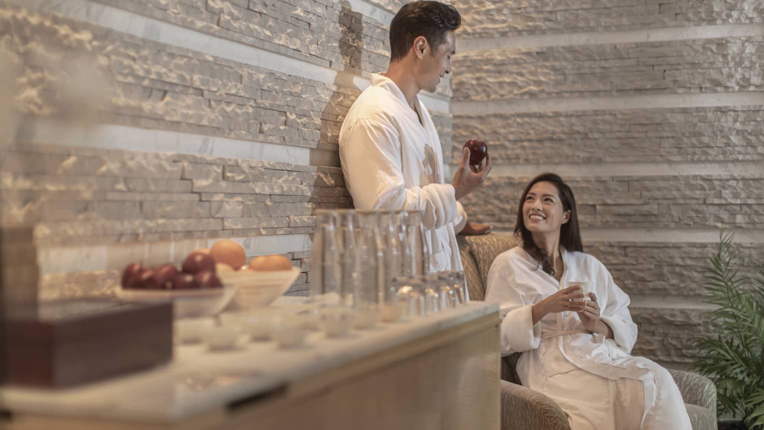 Two people in white robes relax in a spa lounge, one holding an apple. Various fruits and glasses sit on a wooden surface. The atmosphere is calm and inviting.
