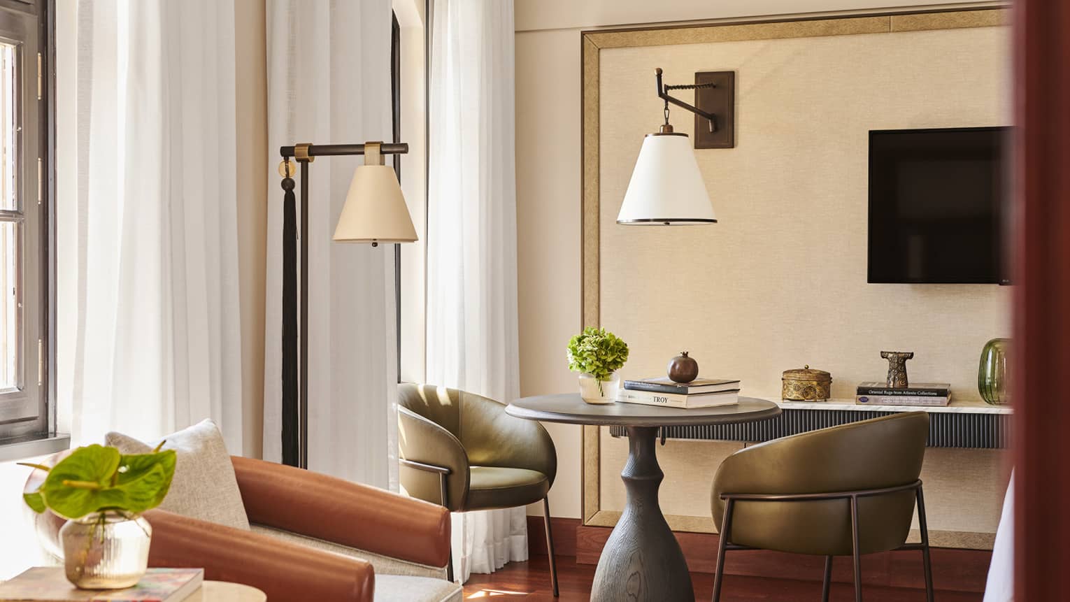 Corner of a hotel room, complete with a round table and two leather chairs, two lamps, an armchair, TV and windows