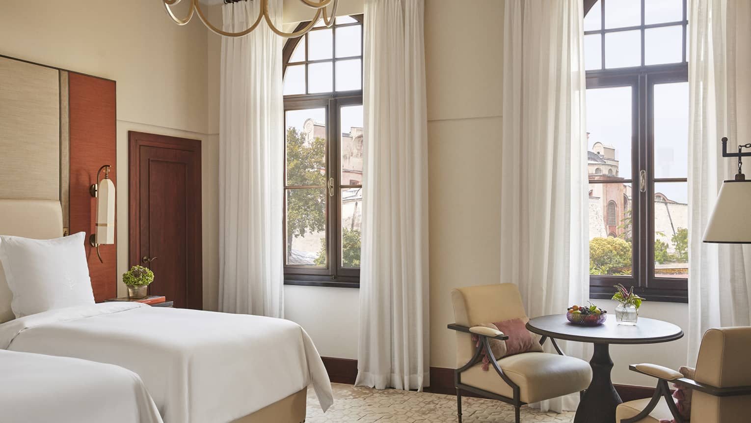 Hotel room with two twin beds, two windows, chandelier and small table with two chairs