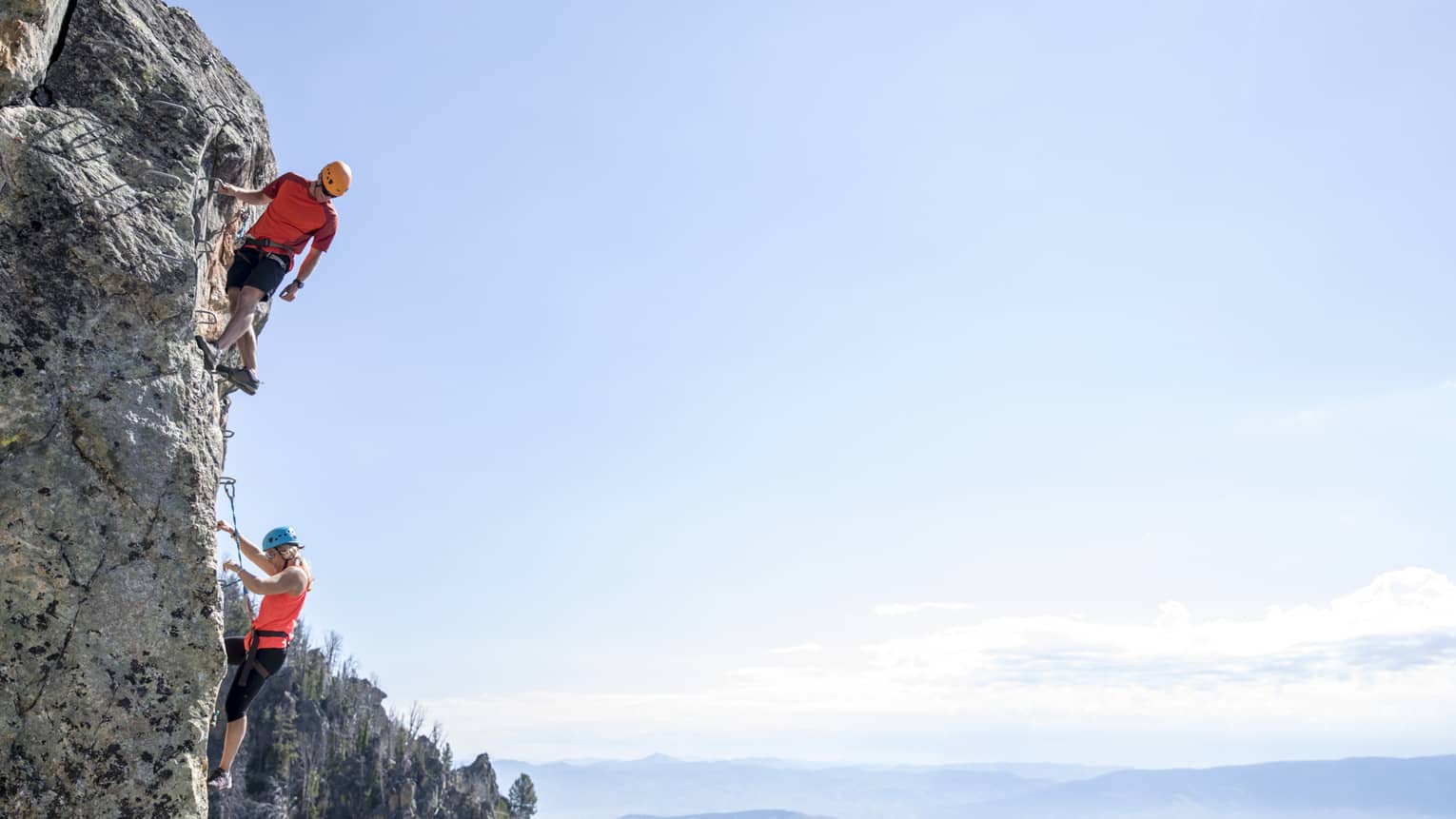 Man and woman climb steep cliff face with view of mountains in far distance 
