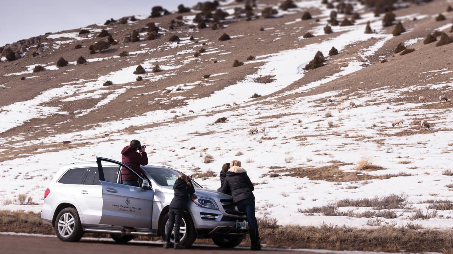 A group of people standing around a small SUV looking at a mountain with snow on it.