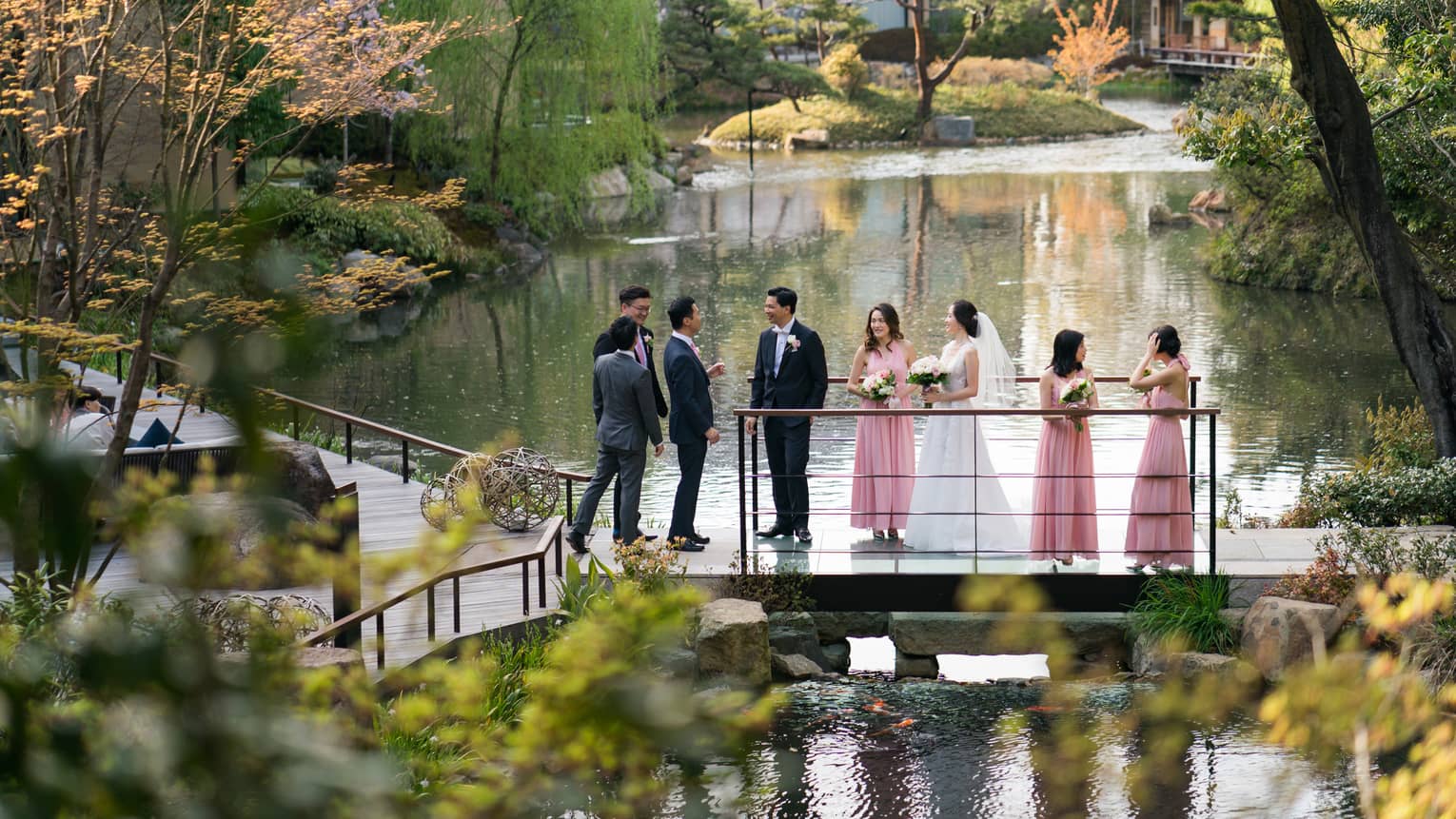 Bride, groom and wedding party stand on small bridge over pond