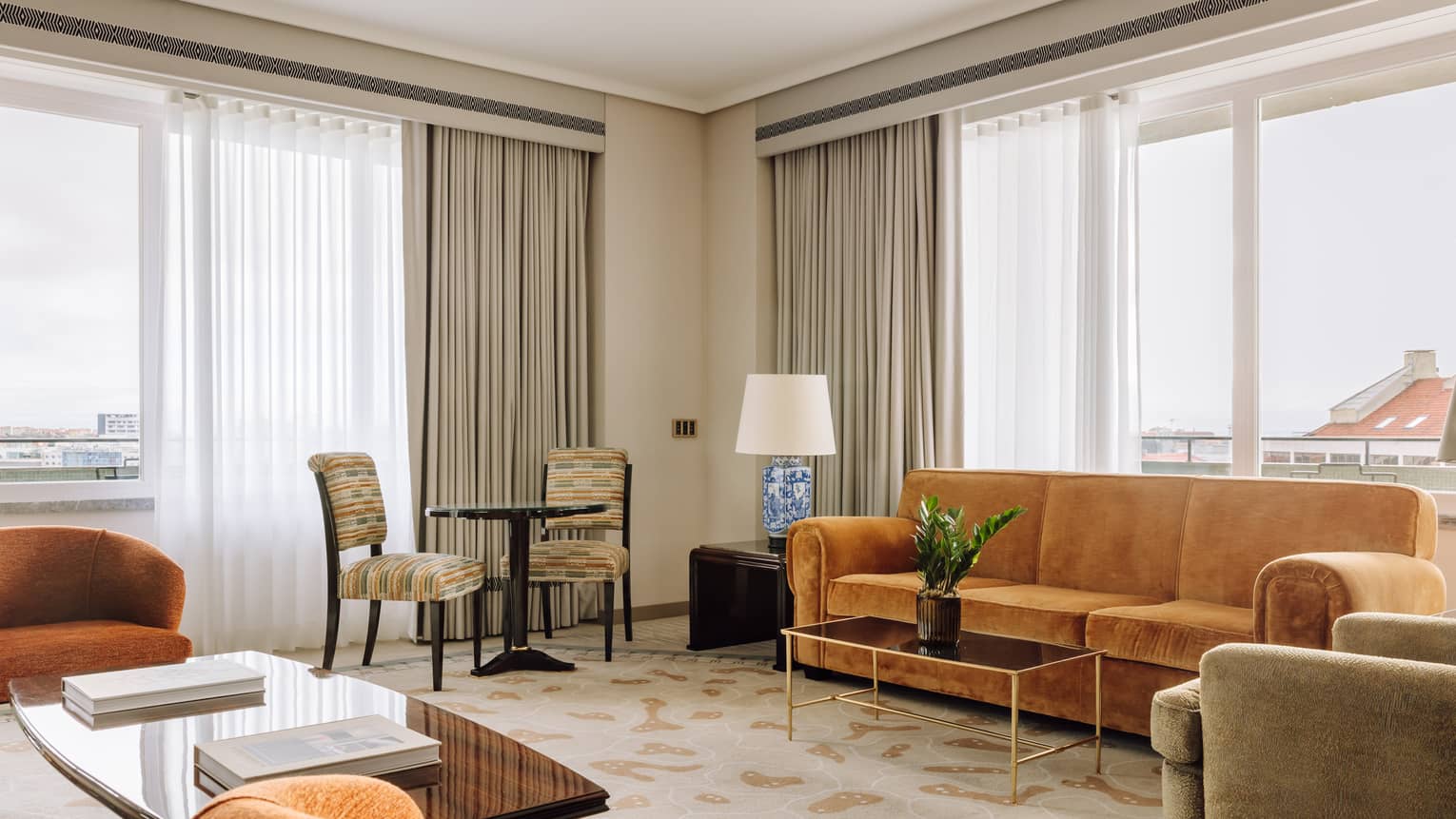 Bright hotel suite living area with sofas, armchairs, a coffee table and a small dining set near large windows with sheer curtains.