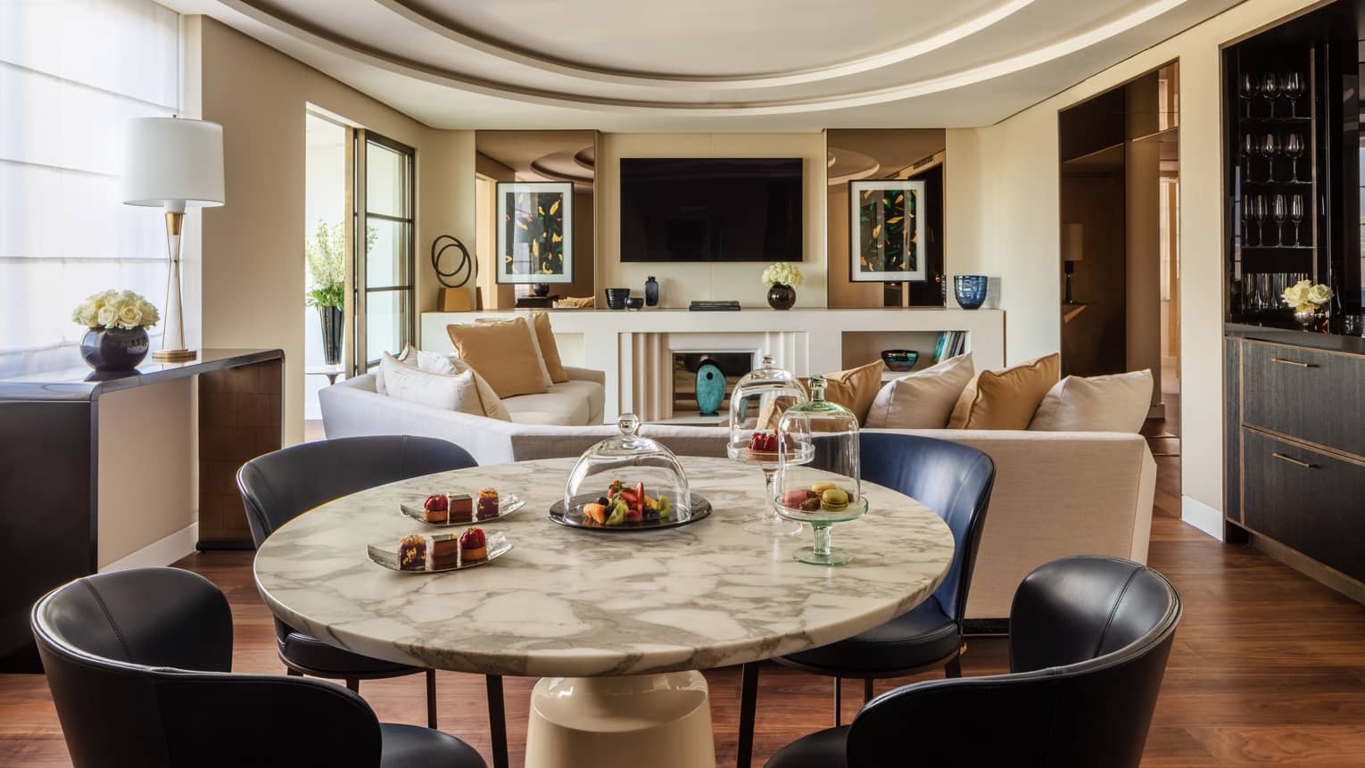 A suite with a round stone table with black chairs, a beige sofa, a television and large windows.