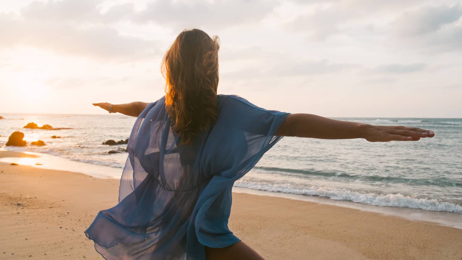 Woman wearing sheer beach cover kneels with arms outstretched in yoga pose on sandy beach by water