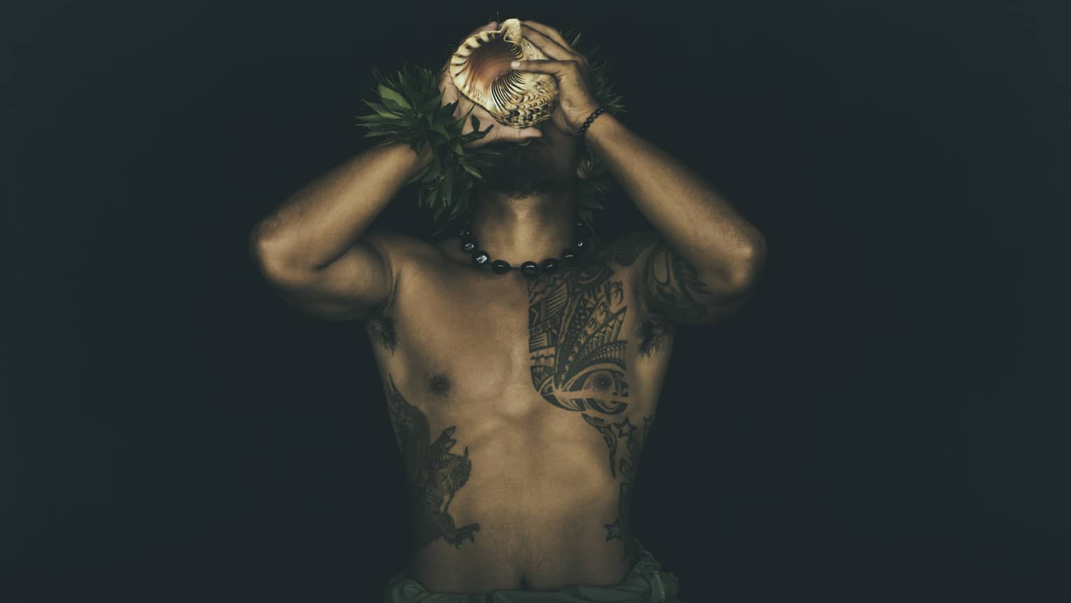 Shirtless tattooed Kānaka Maoli man plays traditional conch shell like a trumpet in the night
