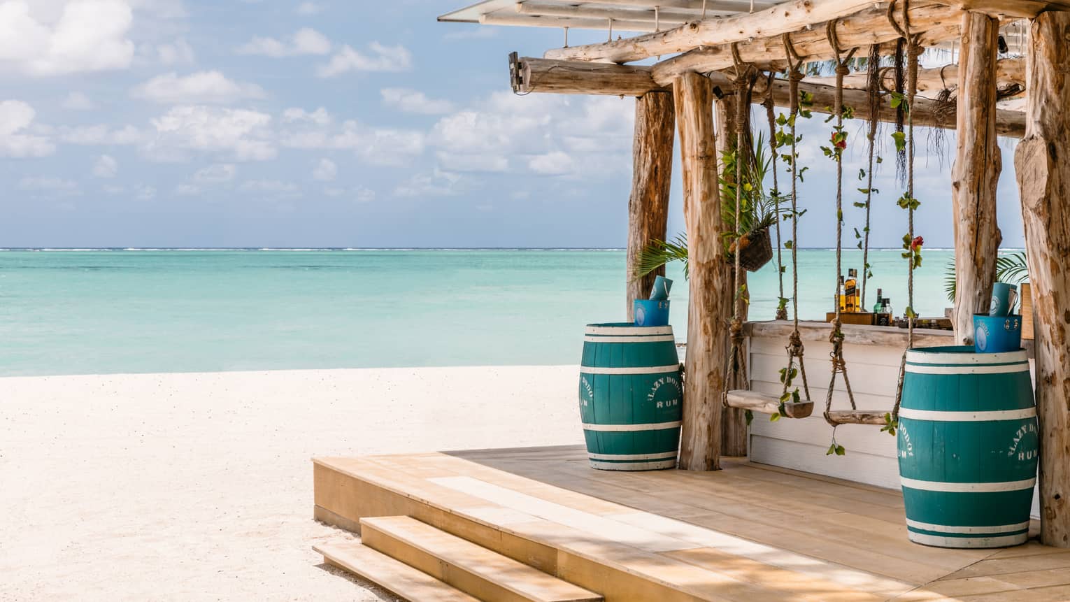 Wooden bar with pergola, two blue barrels, two swings on beach, aqua views, cloud dotted blue sky