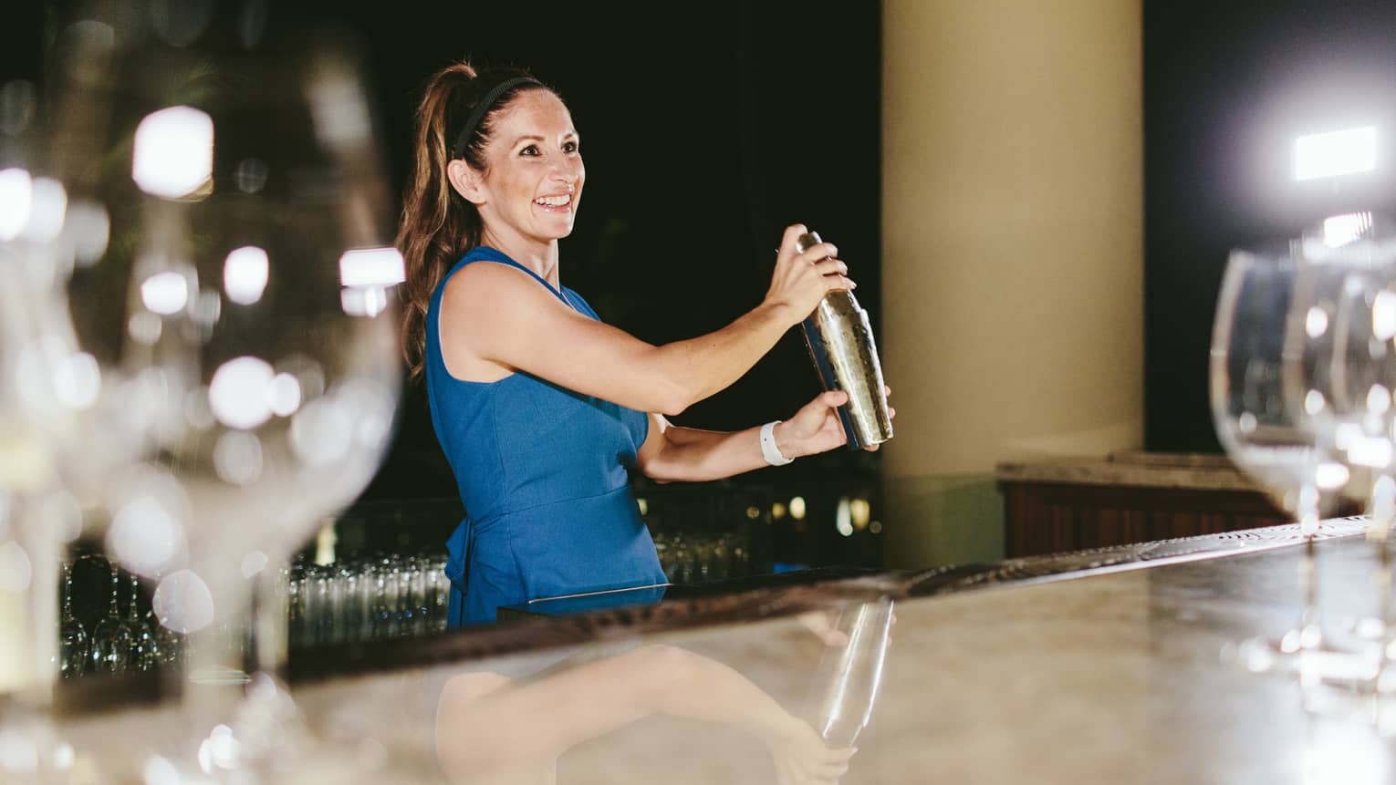 Wine goblets sparkle atop an outdoor bar in the foreground, as a smiling bartender shakes a cocktail beneath a jet-black sky.
