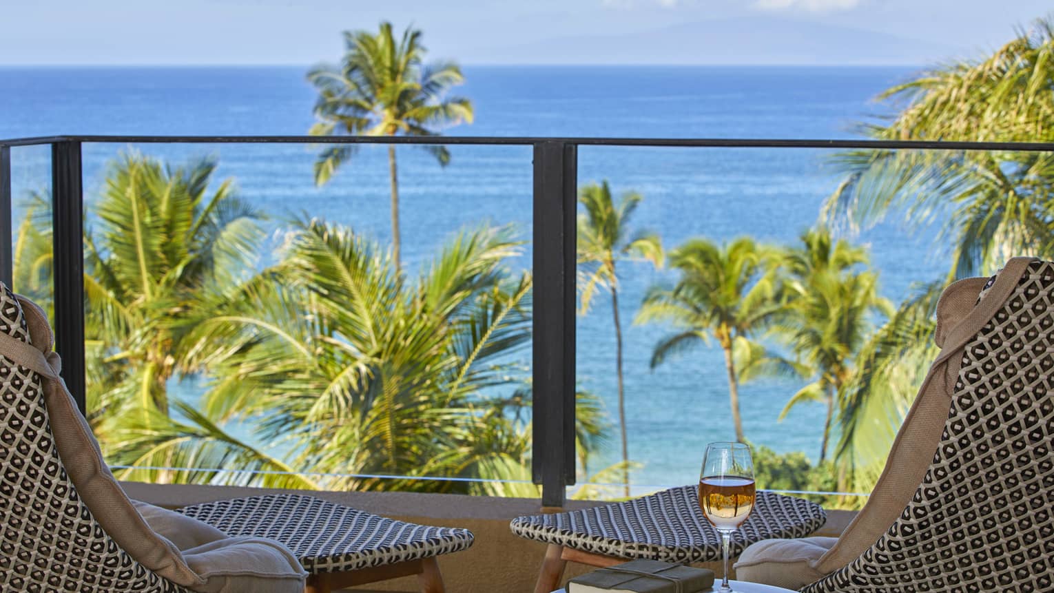 Balcony with two patterned chairs, ocean view palm trees and a table with a book and glass of wine