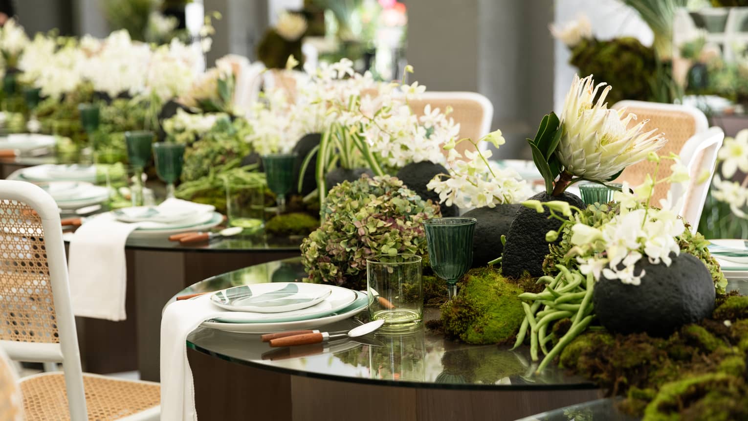 A row of round tables with chairs in a bright event space. Tables have place settings and floral centrepieces of white and green.
