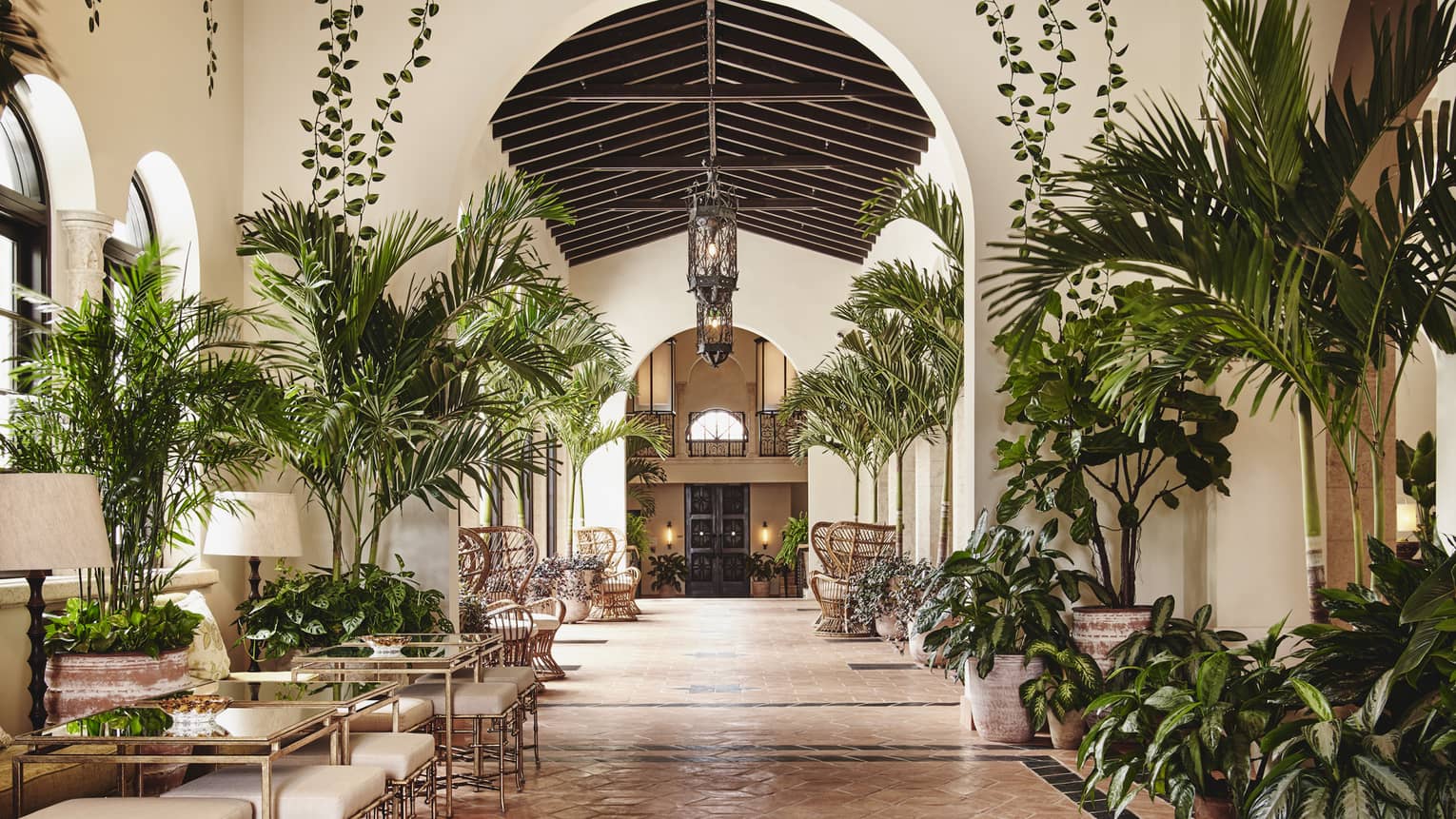 The interior of a lounge area of a hotel with small palm trees, other plants, and tables for people to sit at.