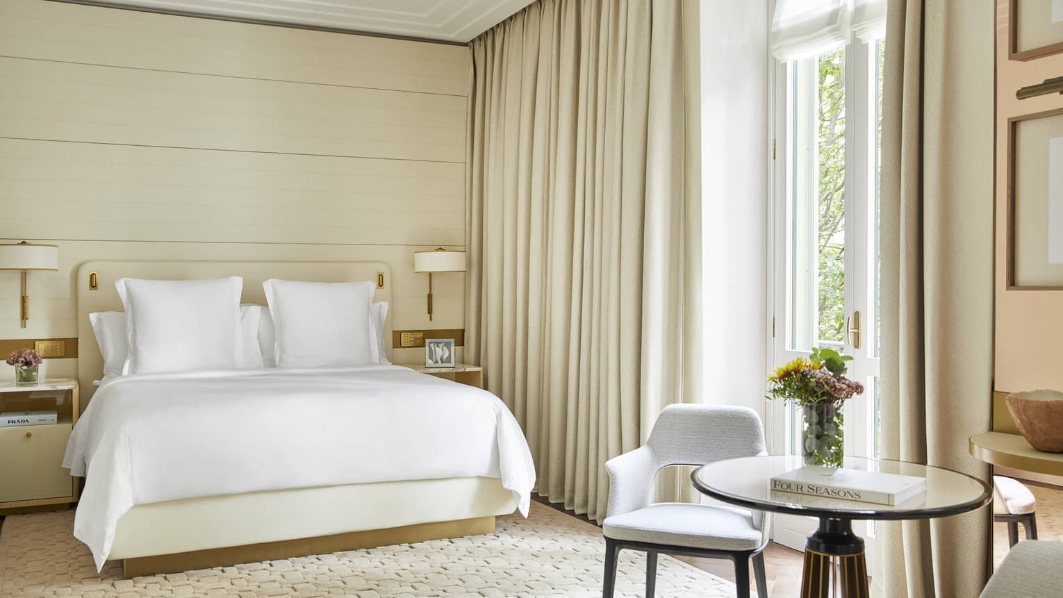Light-filled hotel guest room with king bed, small round table with two chairs and creme-coloured curtains