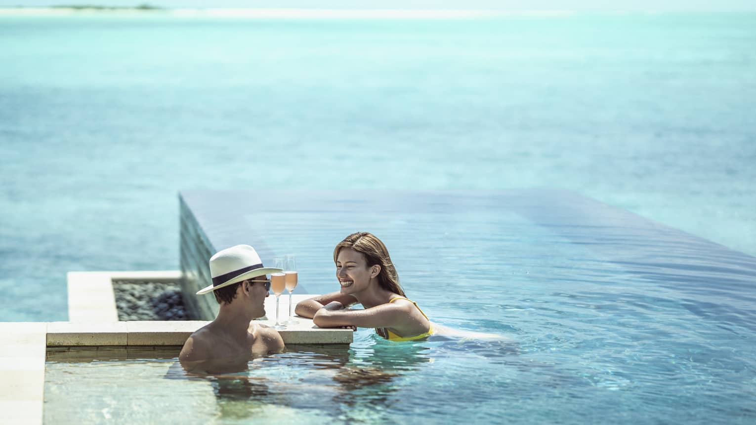 Smiling couple relaxes in infinity pool with glasses of champagne