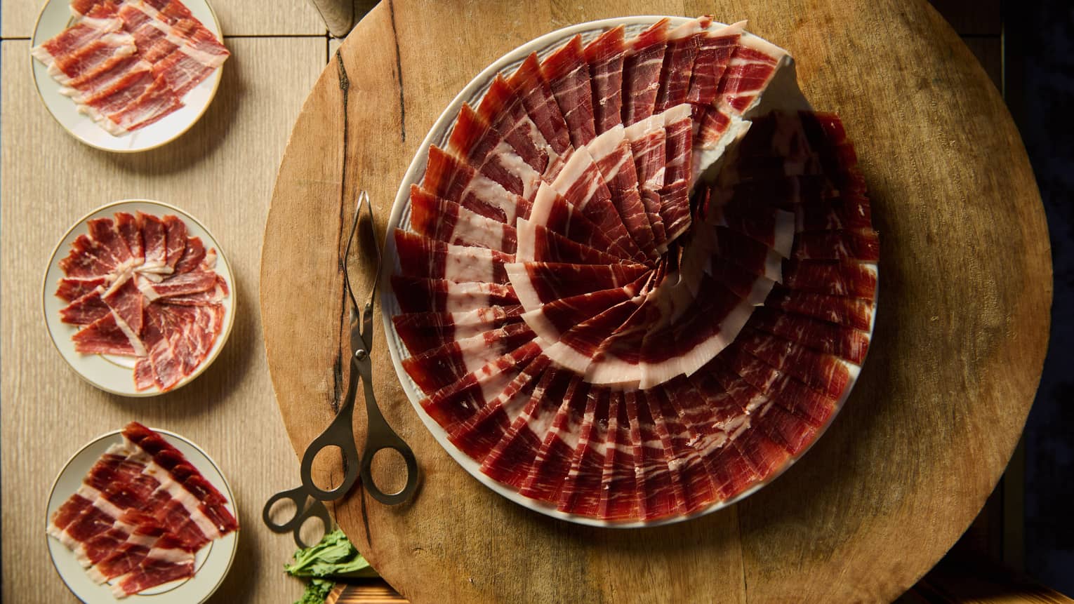 A white plate on a round wood display surface holds thin slices of meat in a decorative spiral pattern. Tongs are next to the plate, and three smaller plates with decorative arrangements of thin-sliced meat are off to the side.