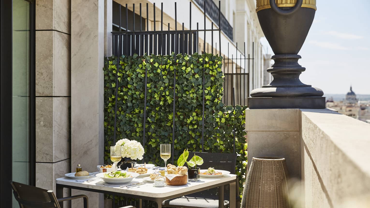 Outdoor dining table on a terrace with plates of food, wine glasses and flowers, set against an ivy-covered fence with city views and decorative urn nearby