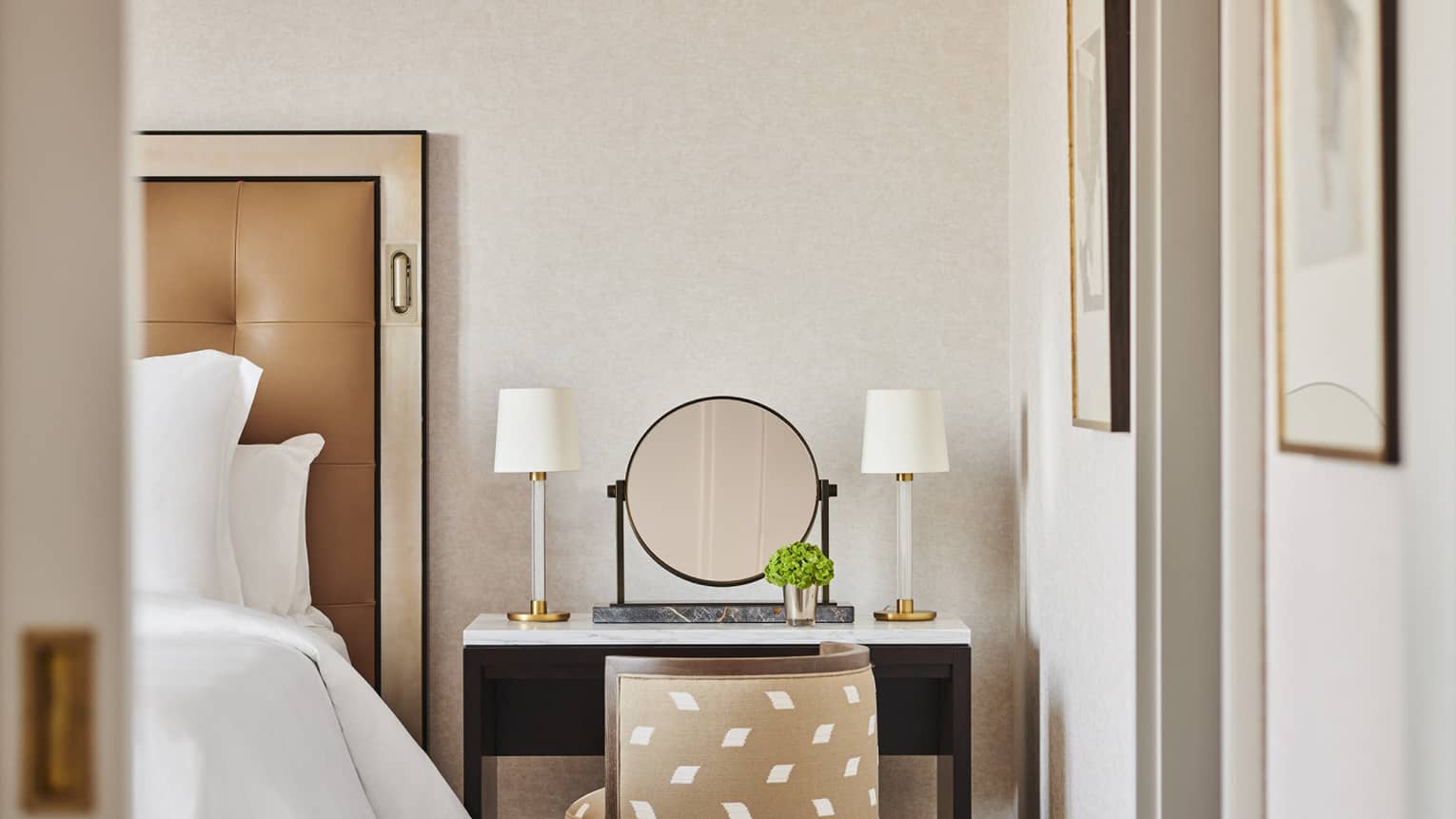 Vanity table beside a king bed in a luxury hotel room featuring a round mirror flanked by two white lamps, a small green plant and a beige patterned chair