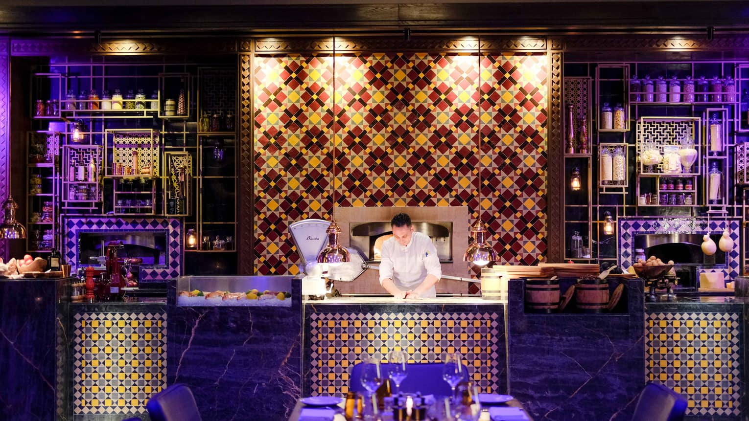 Chef presses dough on counter with colourful tiles, lights, in front of dining table