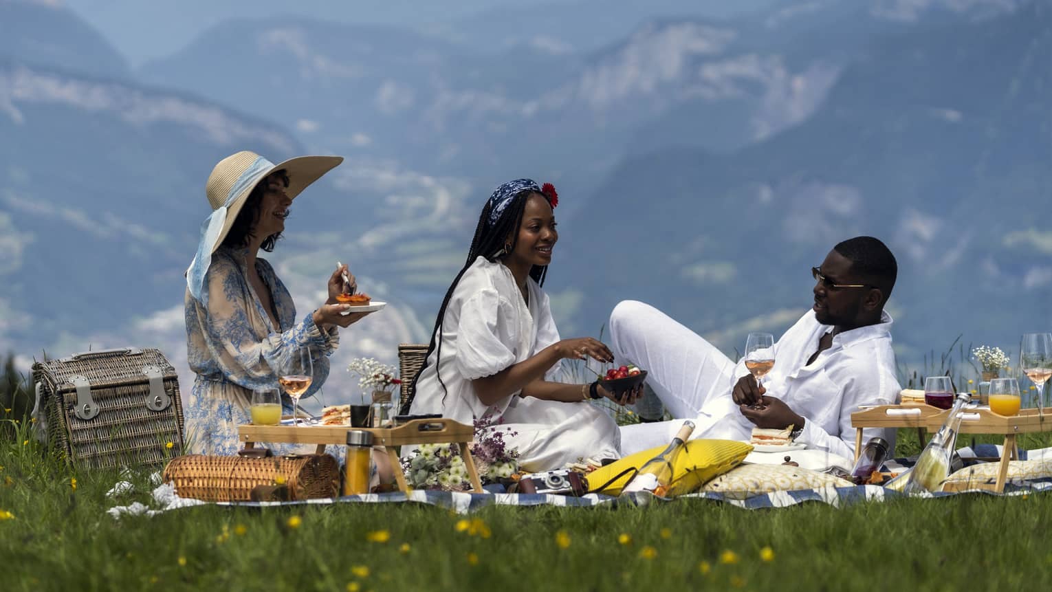 Three friends enjoying a picnic on a meadow with mountains in backdrop