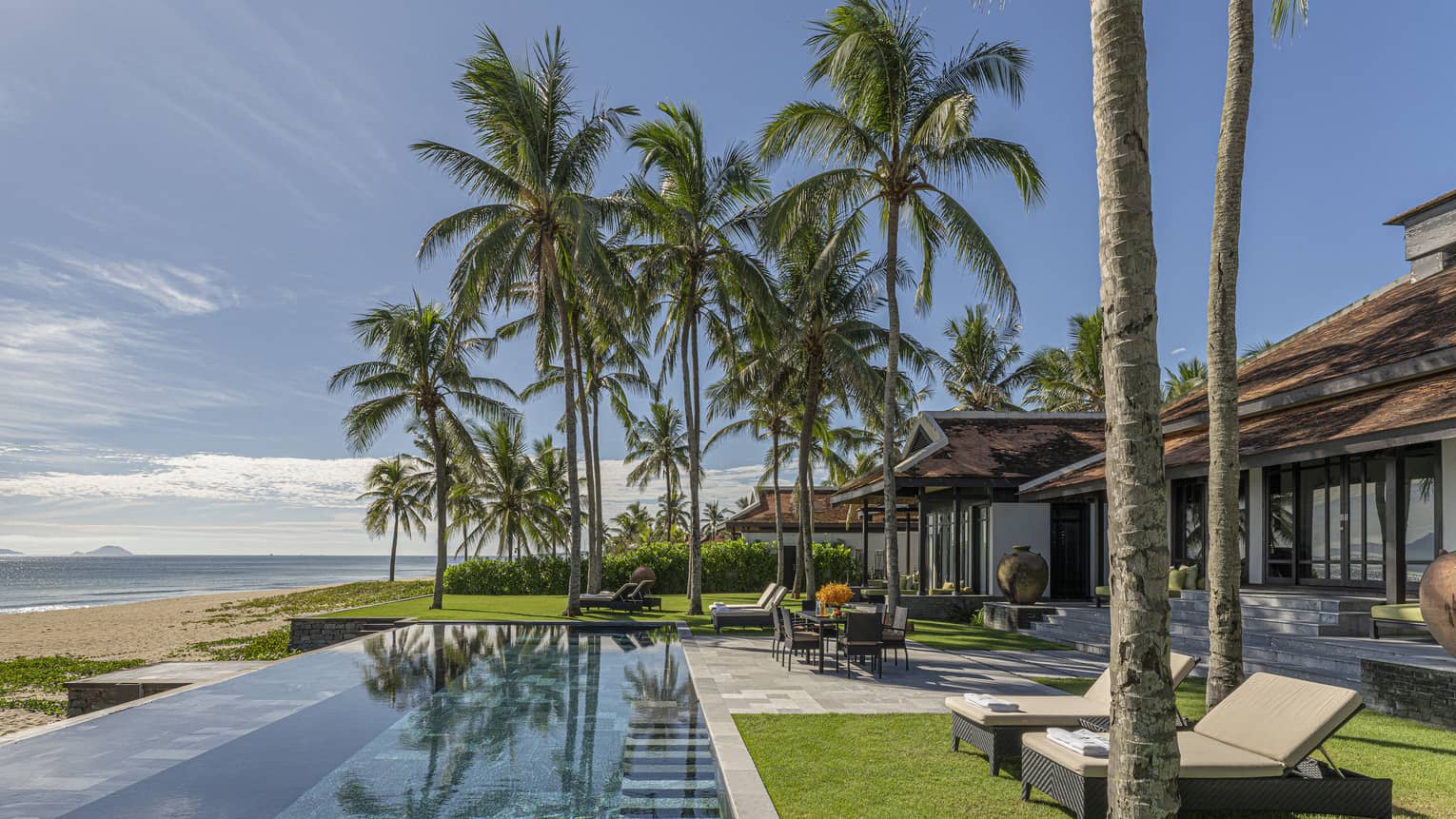 Three Bedroom Beachfront Pool Villa pool area with dining table, lounge chairs, palm trees and blue skies