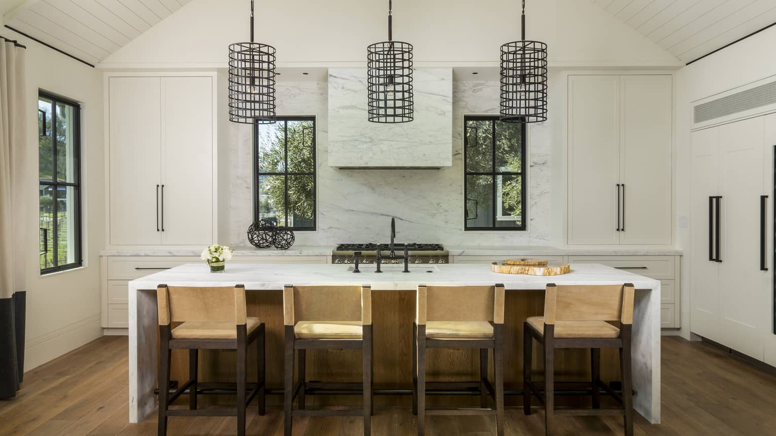 Kitchen with white vaulted ceiling, marble island and accent wall, four tan counter-height chairs and three metal pendant lights