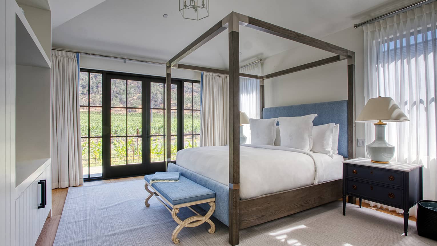 Luxury resort bedroom with a four-poster bed, blue headboard and bench, bedside table with lamp and large windows with drapes overlooking greenery