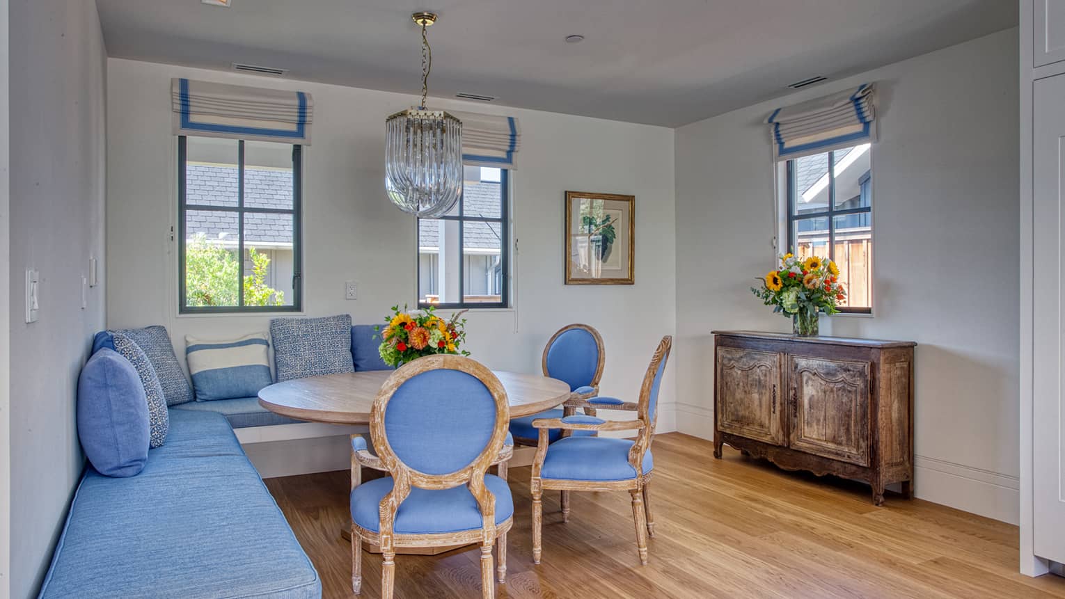 Cozy dining nook with round wooden table, blue upholstered chairs, built-in bench seating with blue cushions, crystal chandelier and rustic wooden cabinet