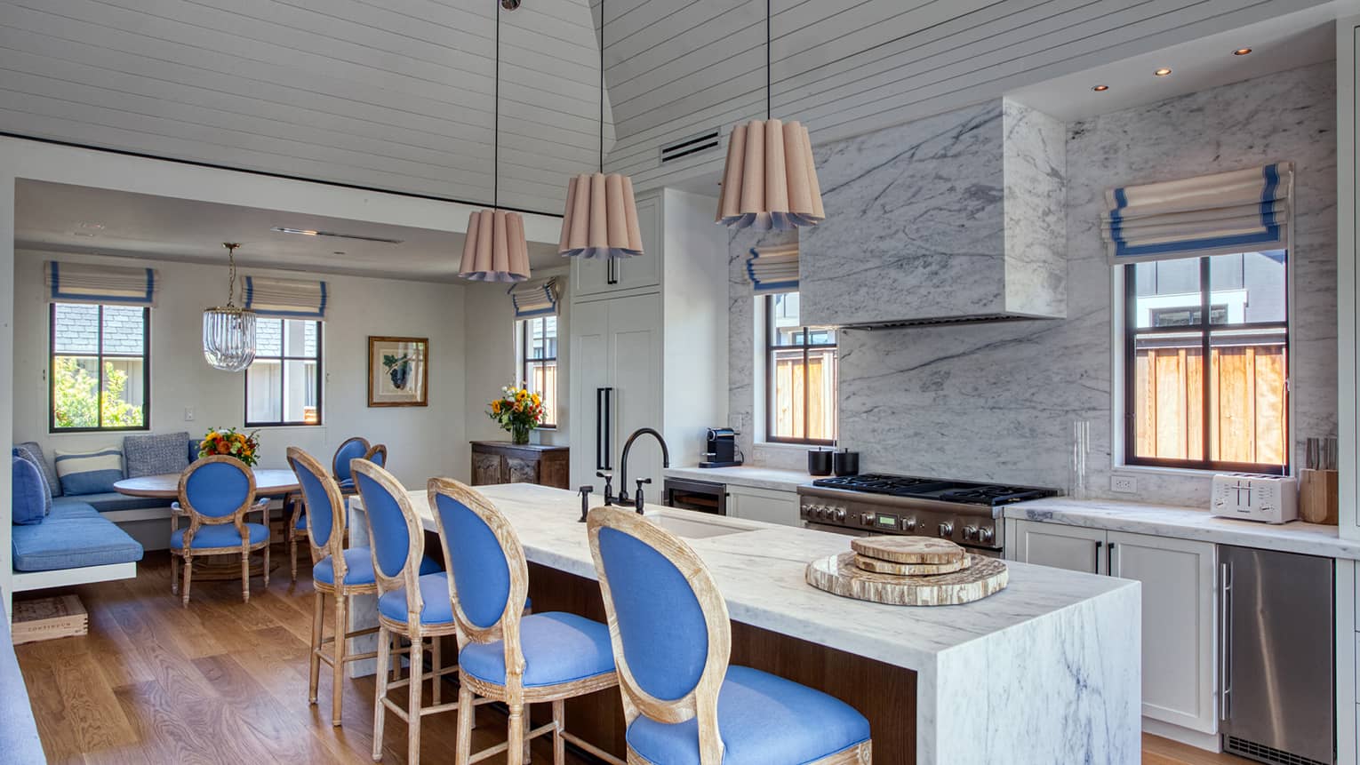 Luxury resort kitchen and dining area with marble countertops, blue chairs, wood finishes, large island with pendant lights, stove and built-in dining nook
