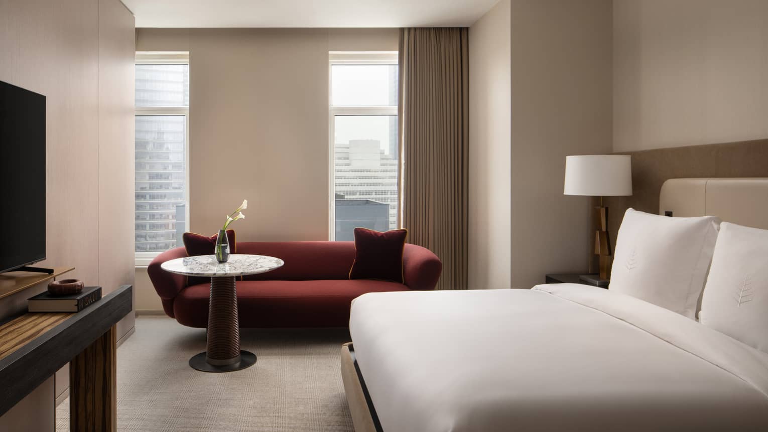 Hotel bedroom with a white bed, red sofa, marble-topped table with a vase of calla lilies, and large windows with beige curtains