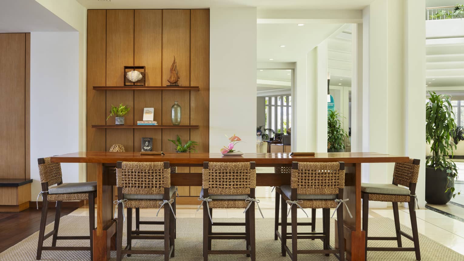 A bright, open interior space with white walls and recessed ceiling lighting includes a long light-brown wood dining table with chairs, a beige area rug, and decorative shelving holding small objects and potted plants.
