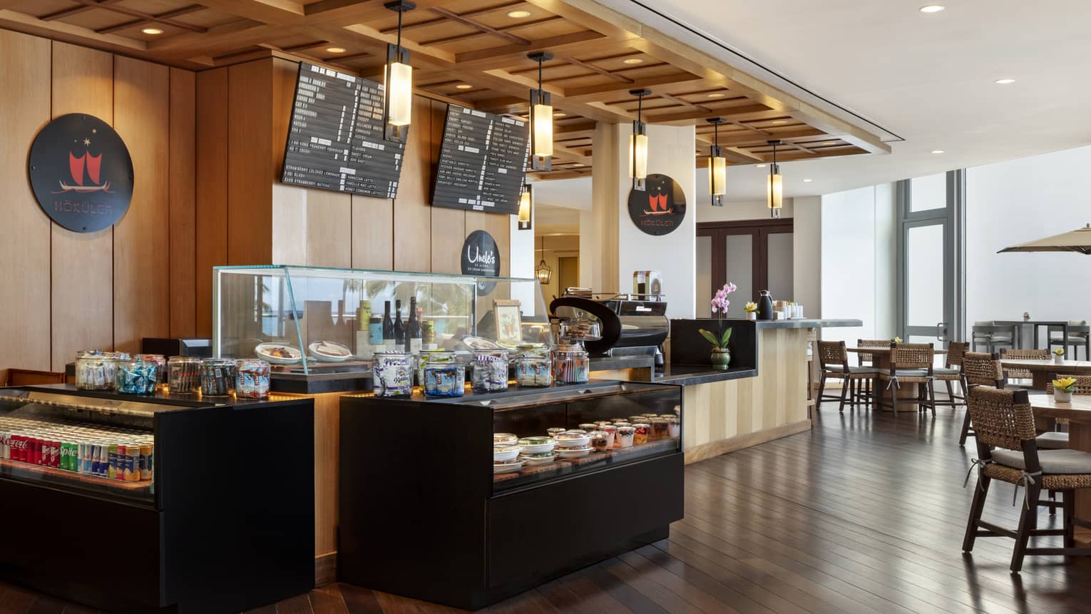 A well-lit restaurant interior space with dark wood floors, warm neutral walls and warm lighting includes large windows, tables and chairs, and a service counter with a display case for food items