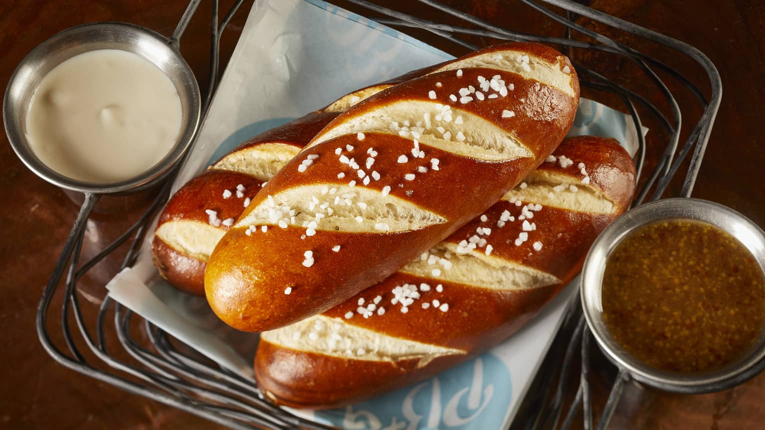 Three large pretzels in a basket with a white and brown sauce in small cups.