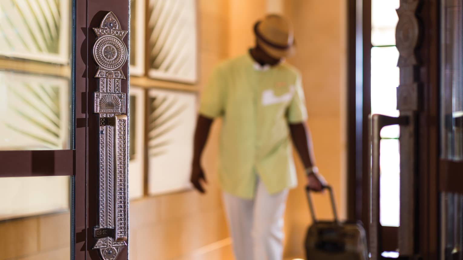 Man with fedora hat wheels suitcase through wooden doors
