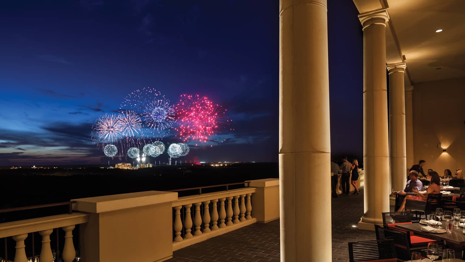 The terrace of a restaurant with tables and people watching fireworks in the distance.