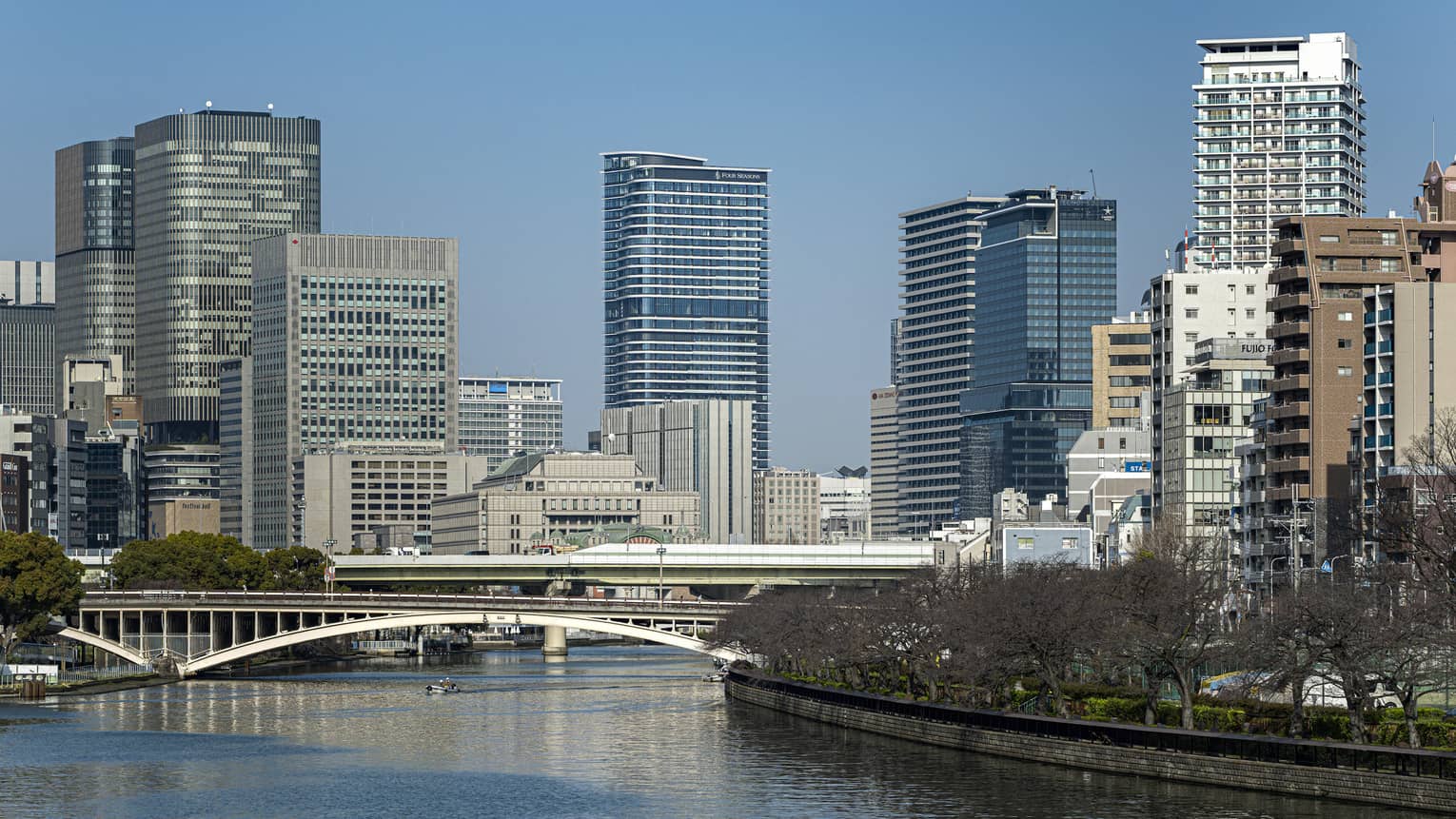Under a clear blue sky, Osaka's modern high-rises loom above two bridges spanning a river lined with dormant cherry blossoms.